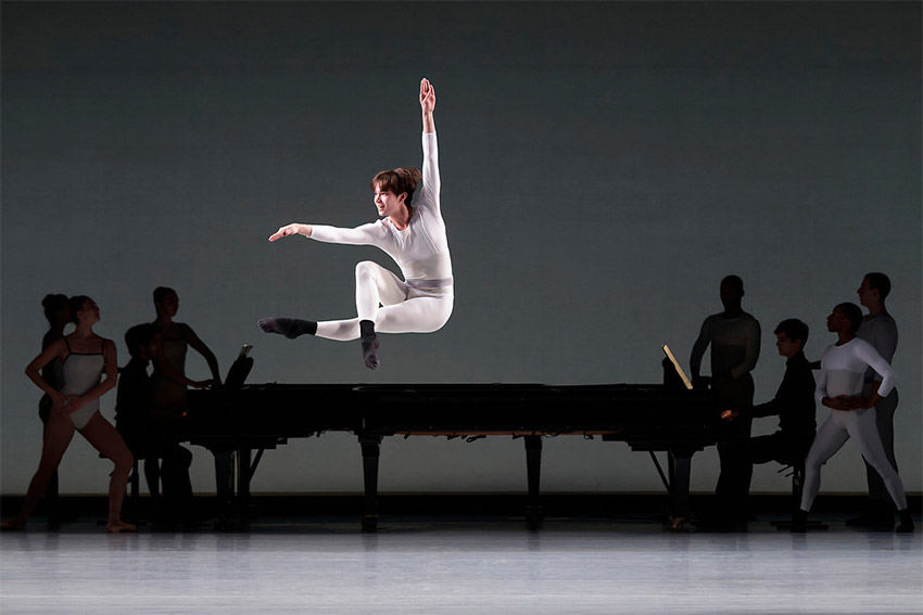 Juilliard dance students. Spotlight on dancer in white leaping over two grand pianos surrounded by other dancers in shadow.