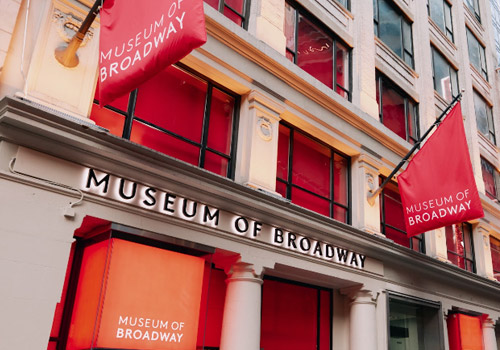 Street view of the Museum of Broadway, a modern building with large red banners displaying its name. Pedestrians walk along the sidewalk and cars are parked on the street in an urban setting with tall buildings.