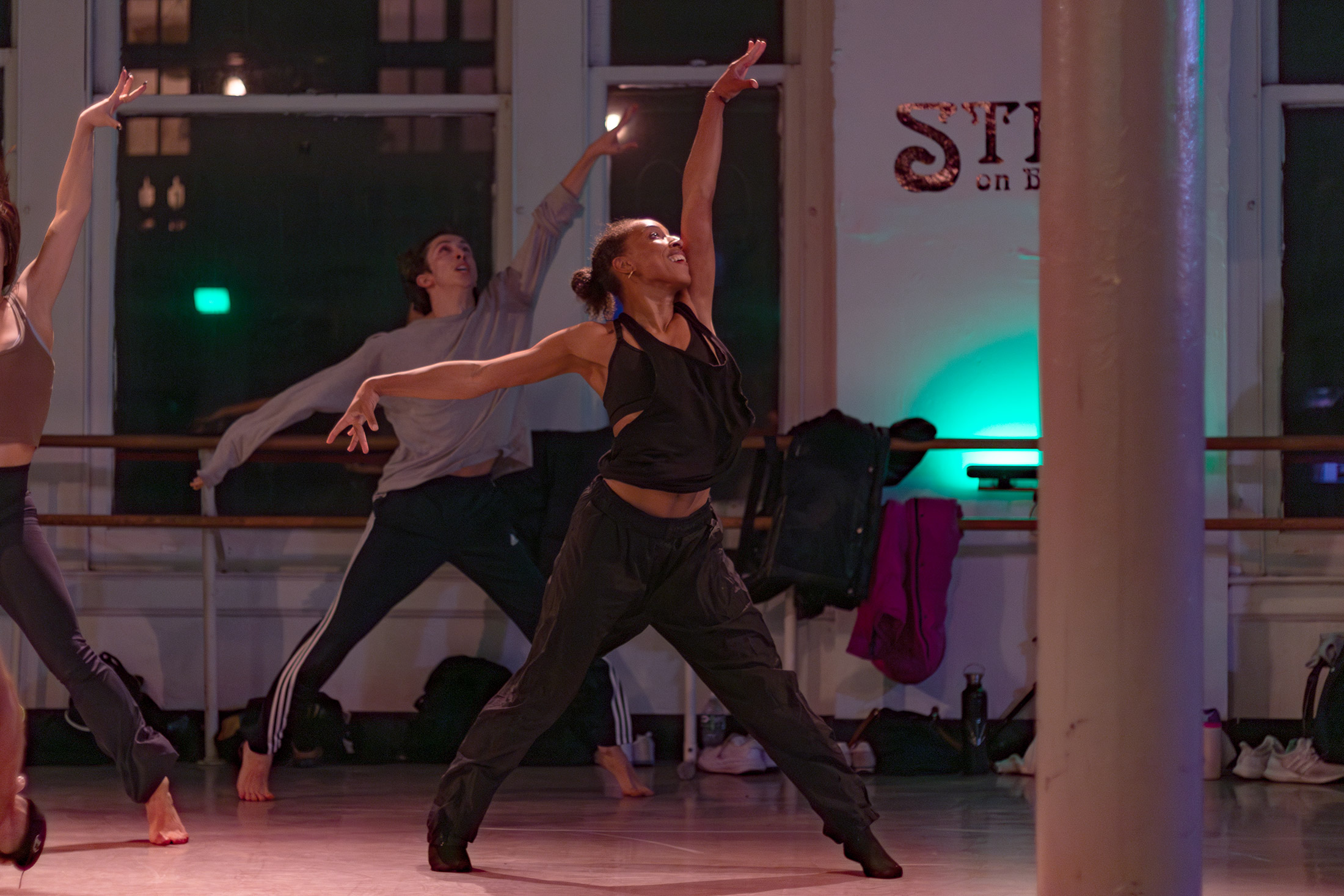 A group of dancers in a studio, with one in the foreground striking an expressive pose with outstretched arms. The setting is softly lit, and there are large windows and walls with Studio partially visible in the background.