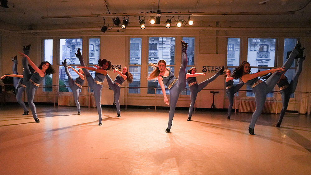 Dancers in matching blue outfits perform a synchronized routine in a studio with large windows. Each dancer is executing a high kick, showcasing their flexibility and coordination. The room is softly lit, and a cityscape is visible outside the windows.