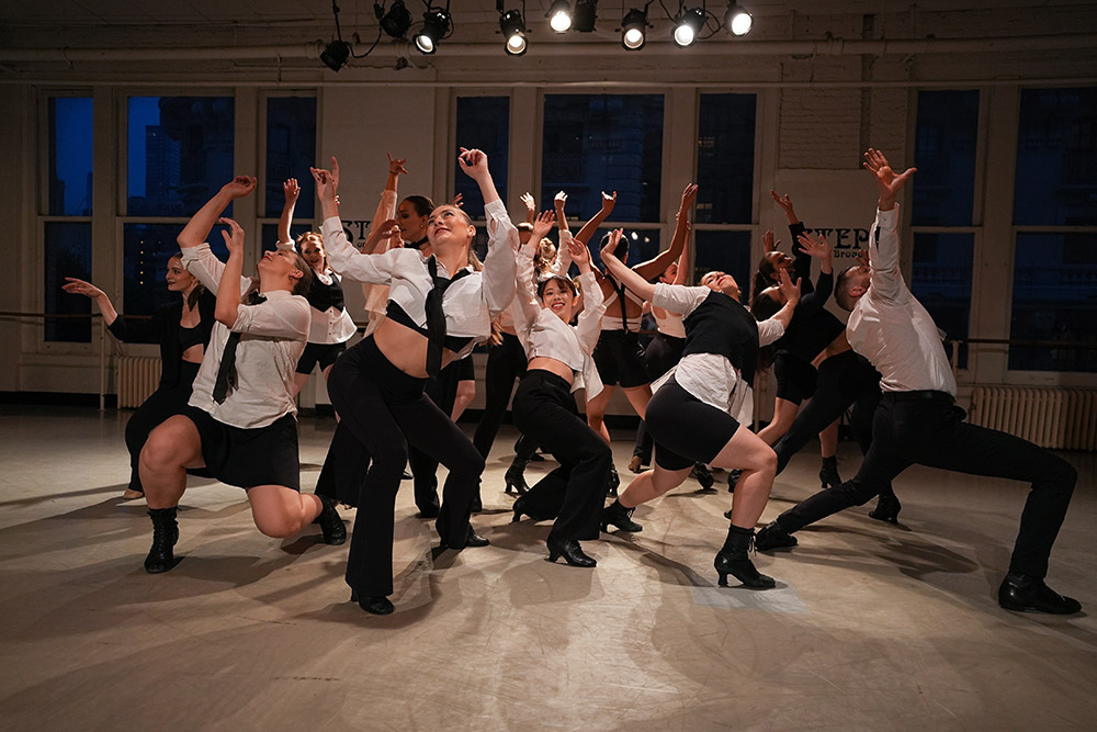 A group of dancers in dynamic poses perform a contemporary routine in a studio. They wear coordinated black and white outfits and are energetically extending their arms and legs. Large windows reveal an urban backdrop.