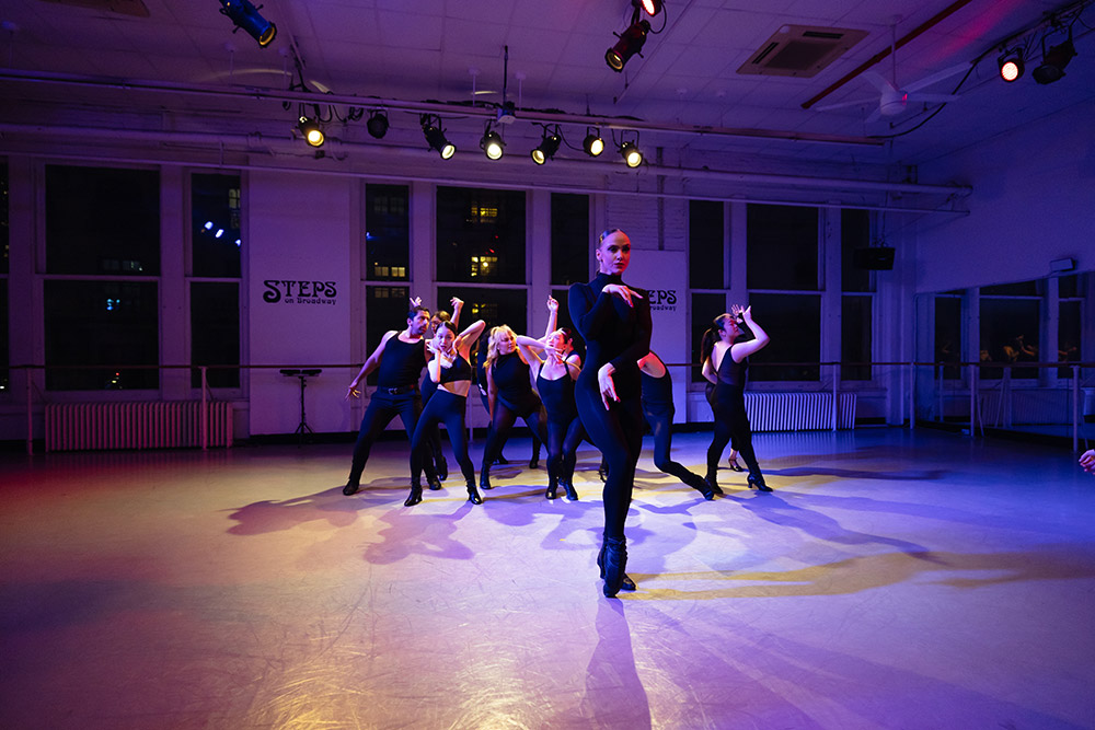 A group of dancers in black outfits performs energetically in a studio with STEPS signage on the walls. Stage lights illuminate the dancers as they strike dynamic poses against a background of large windows.