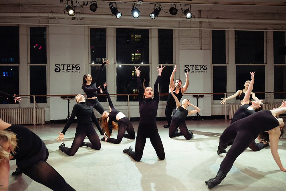 A group of dancers in black outfits perform an expressive routine on a studio floor with large windows in the background. They are striking dynamic poses under bright overhead lights, with the words Steps on Broadway visible on the walls.