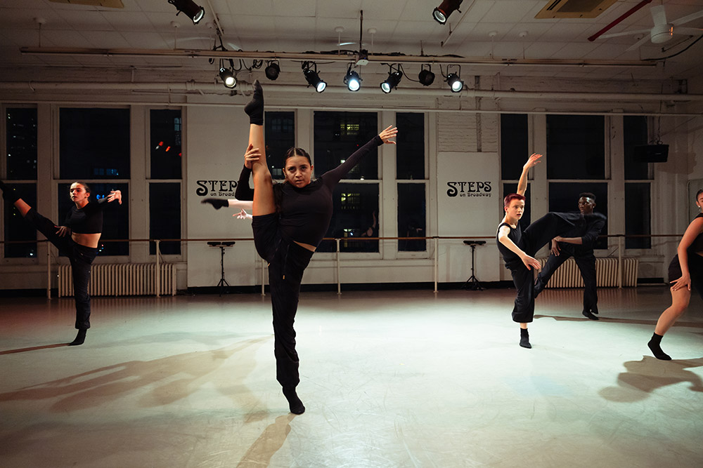 Dancers in a studio perform synchronized moves. One dancer in the foreground lifts a leg high while others execute dynamic poses in the background. The room is dimly lit, with large windows and STEPS on the wall.