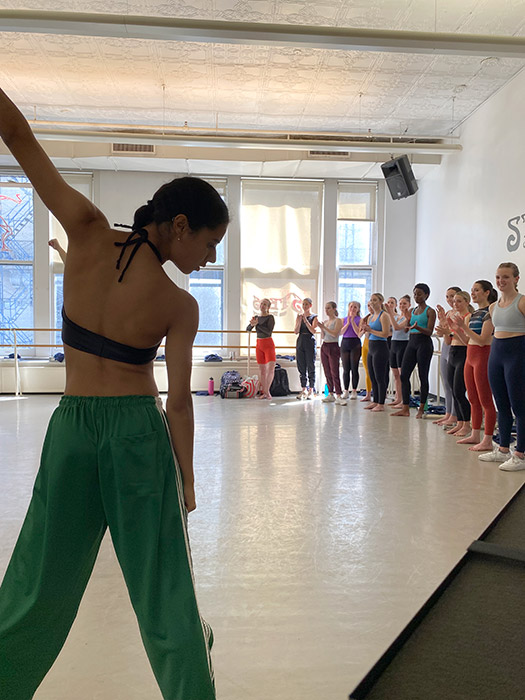 Steps Summer Study students in the dance studio one in the foreground with her left arm up, many others lined up on the right wall