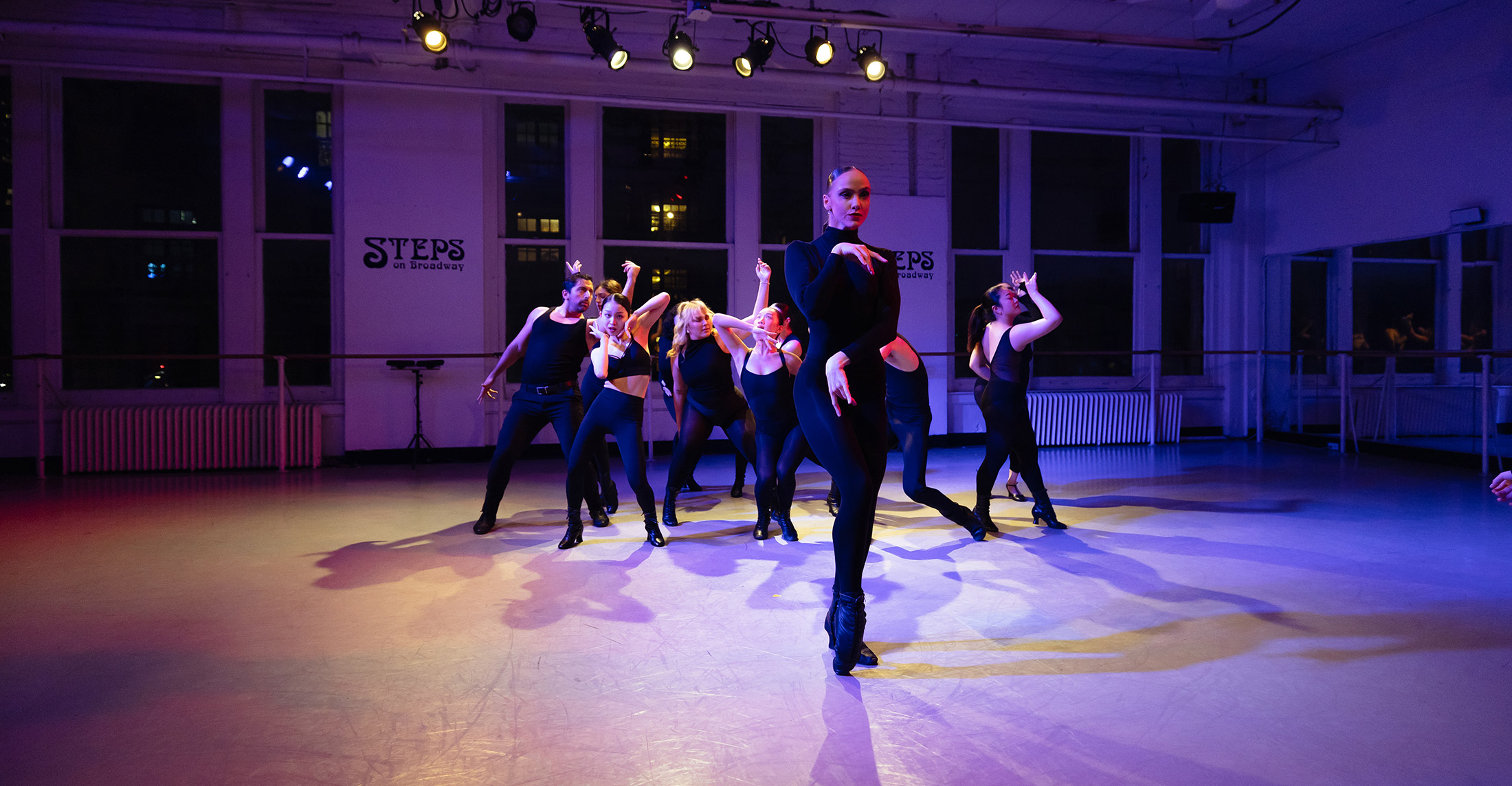 A group of dancers in black outfits perform energetically in a dimly lit dance studio with large windows. Stage lights illuminate the scene, highlighting their dynamic movements. A dancer in the foreground leads the formation.