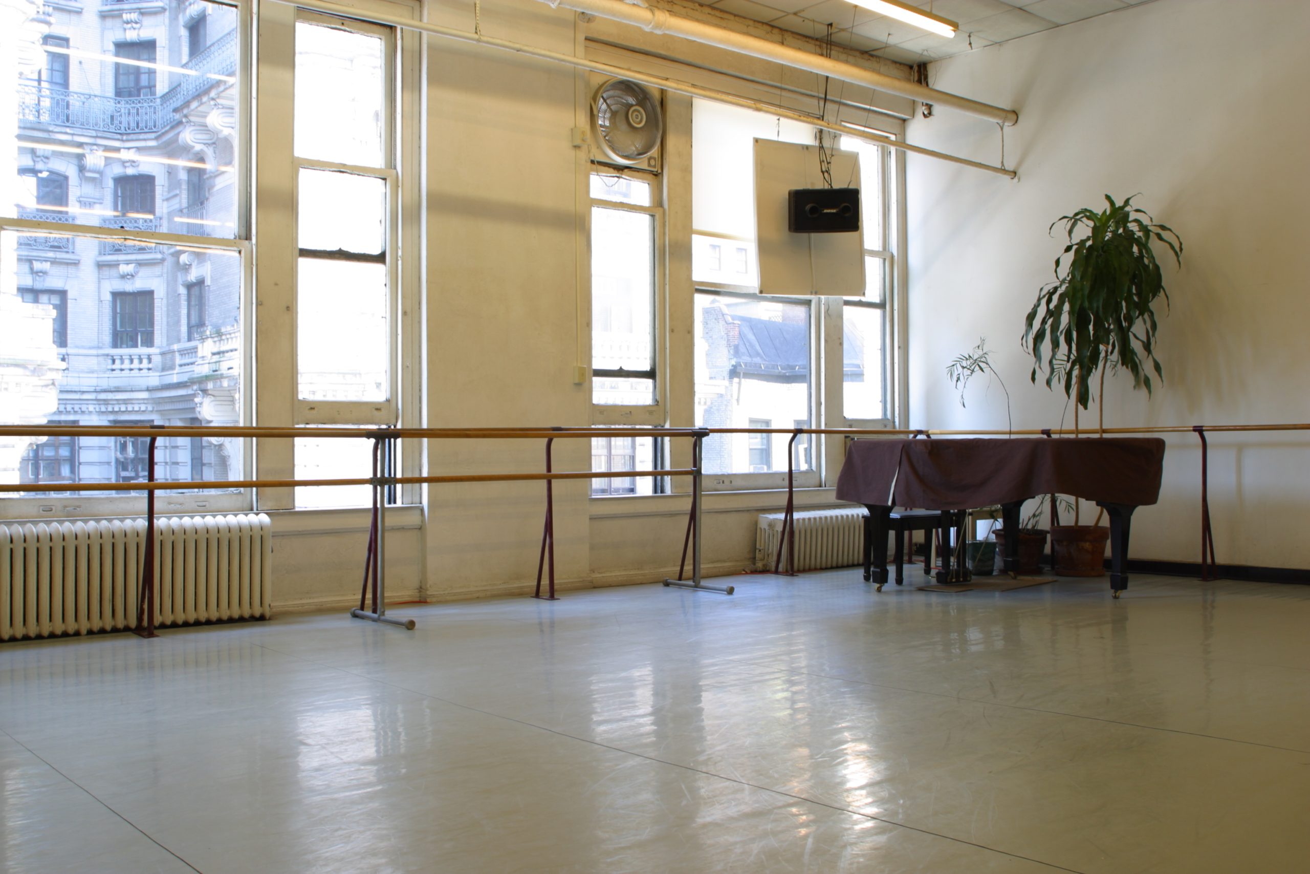 Steps Studio 2 - the corner of a dance studio with a ballet barre in front of soaring windows looking out on NYC with a grand piano and a potted tree in the corner