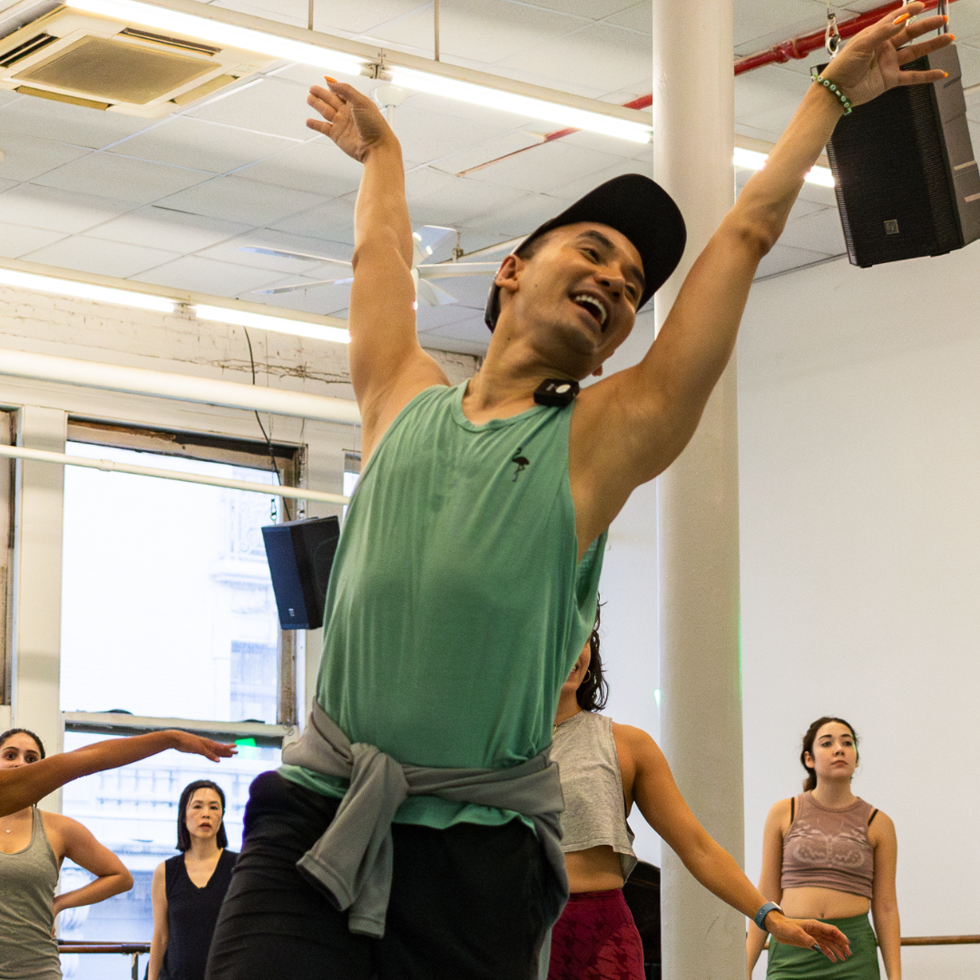 A dancer in a green tank top energetically moves with one arm raised in a studio. Several other people are visible in the background, also participating in the class. The room has large windows and bright lighting.