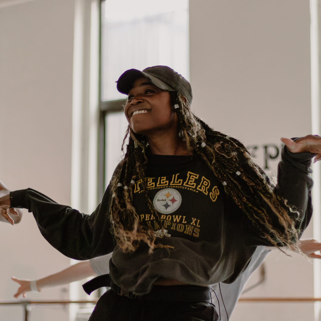 A person wearing a cap and a Steelers sweatshirt is smiling and teaching dance in Steps Black Voices in Dance series. Their hair is styled in long braids, and their arms are outstretched. The background is softly blurred, with light coming in through a window.