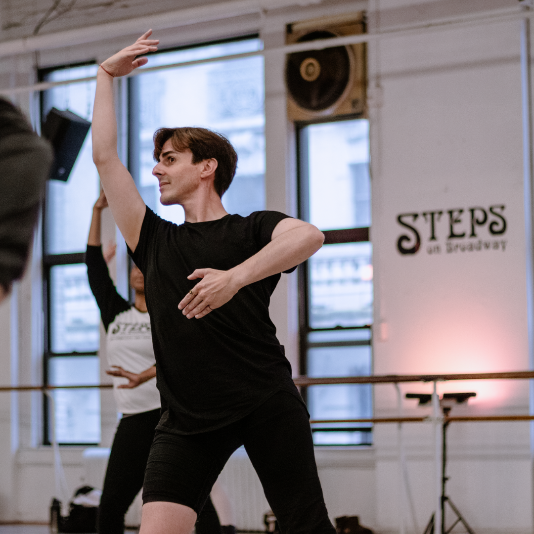 A dancer in a black outfit strikes a pose in a dance studio with large windows. Another person is visible behind him, partially obscured. The studio wall has a Steps on Broadway sign.