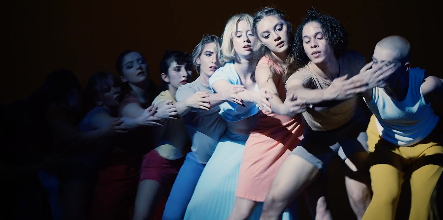 A tight group of UNSCA dance students on a dark stage lined up behind one another with arms reaching forward
